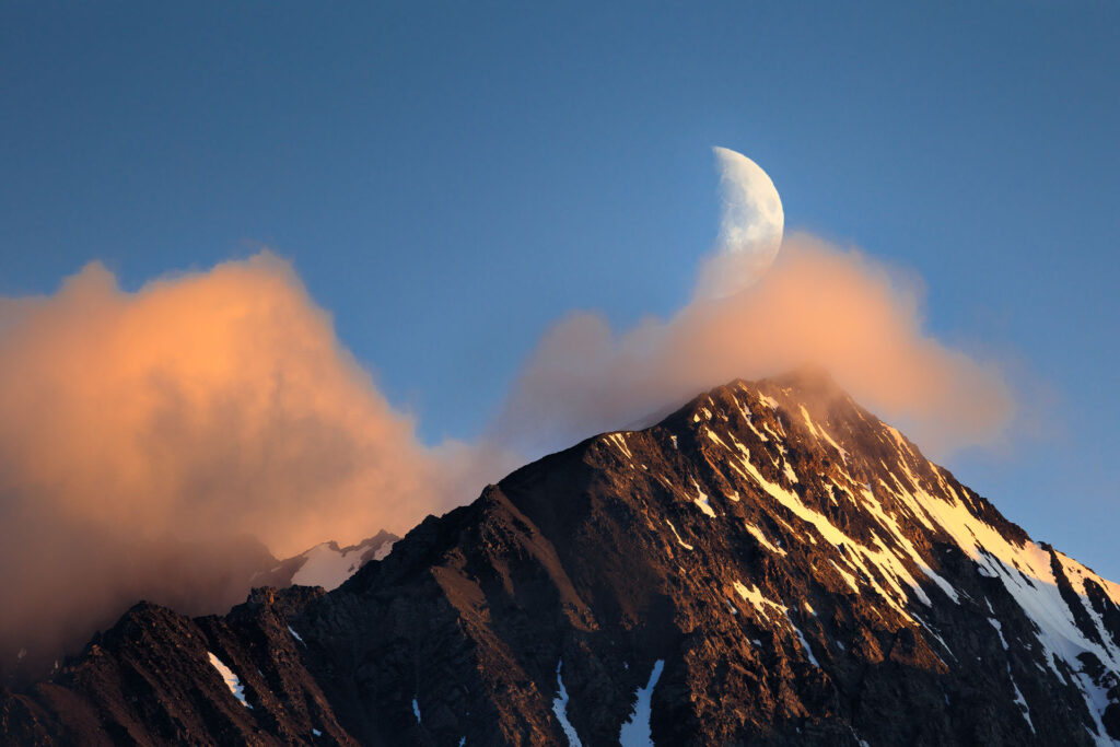 moon sets over sunset-lit clouds and mountain peaks