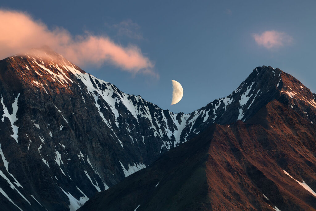 moon sets while sunset-lit clouds reach out over mountain peaks